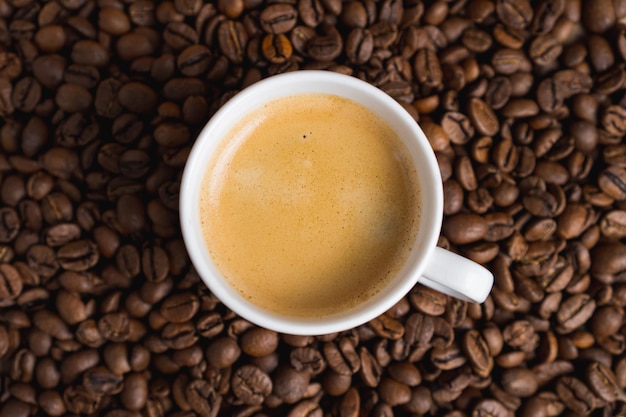 Small white coffee cup with espresso crema on roasted beans background Selective focus