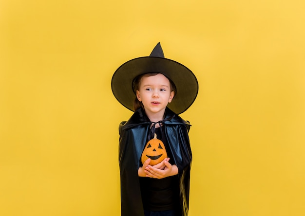 A small witch in a hat holds a painted pumpkin on yellow