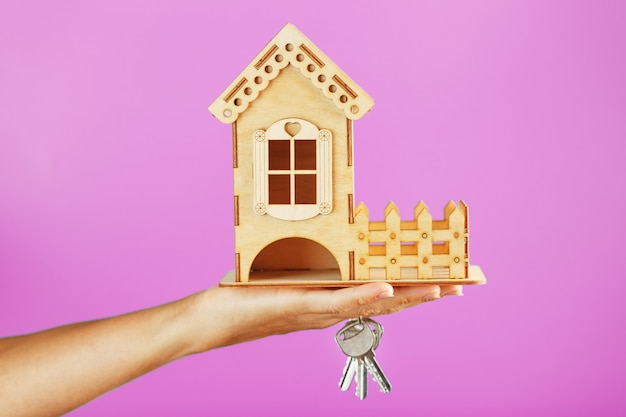 Foto una piccola casa di legno con le chiavi in mano su uno sfondo rosa.