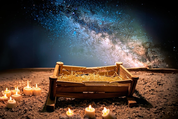 Small wooden manger filled with straw on snow and candles around and a blue sky