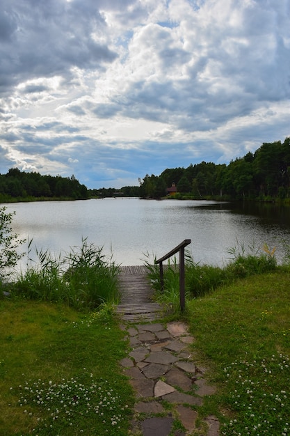 Small wooden pier with a railing on the shore of the lake and a stone path against the blue sky with clouds