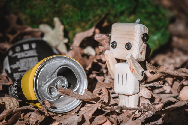 Small wooden robot next to a metal can in forest concept of protection and environment pollution