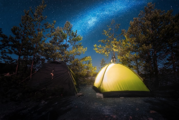 Small yellow glowing tent in the middle of the forest at night with starry sky.