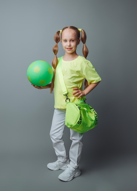 A small young girl with a green sword in her hand smiles at the camera isolated on a gray background a little athlete with a ball children's sports
