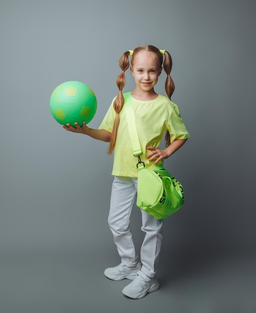 手に緑の剣を持つ小さな若い女の子は、灰色の背景に分離されたカメラに微笑みます ボールを持つ小さなアスリート 子供のスポーツ