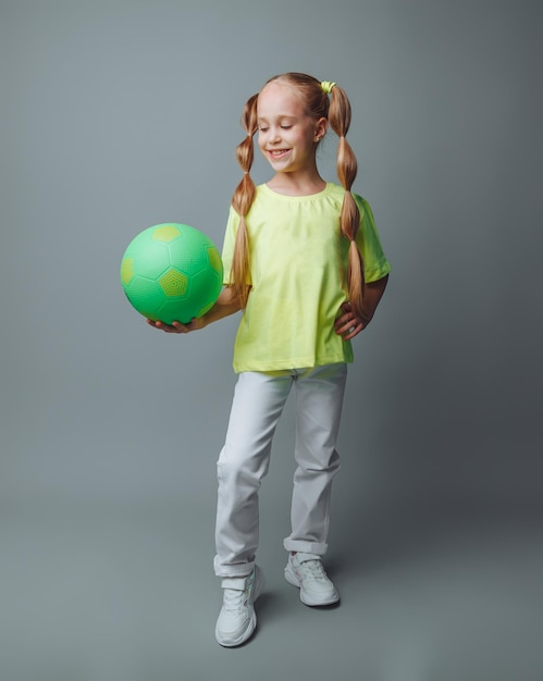 A small young girl with a green sword in her hand smiles at the camera isolated on a gray background a little athlete with a ball children's sports