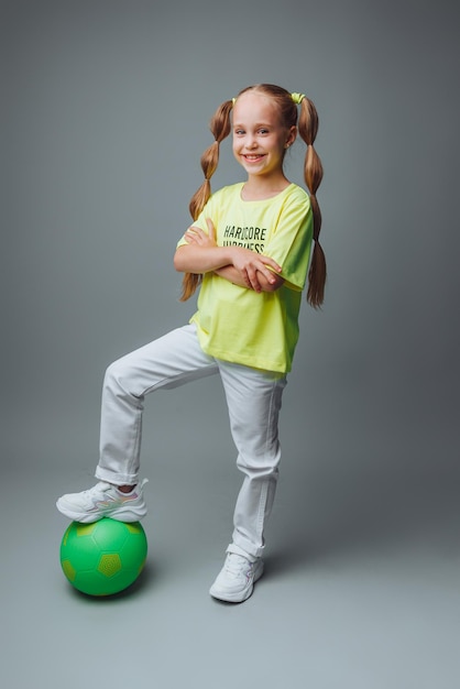 A small young girl with a green sword in her hand smiles at the\
camera isolated on a gray background a little athlete with a ball\
children\'s sports