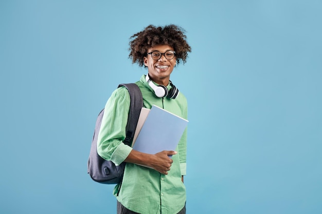 カメラの青いスタジオで笑っているバックパックとノートブックを持つスマートなアフリカ系アメリカ人の学生の男