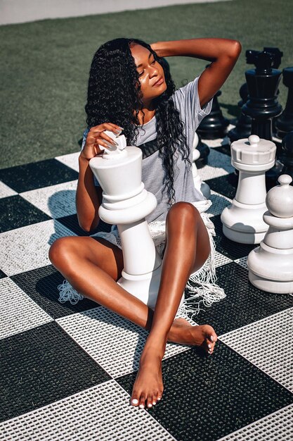 Smart African-American woman playing giant chess as she sits on the board with her legs crossed; glamourous intelligence concept.