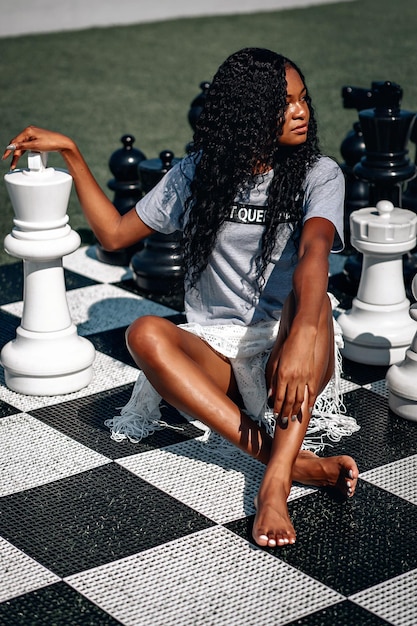 Smart African-American woman playing giant chess as she sits on the board with her legs crossed; glamourous intelligence concept.