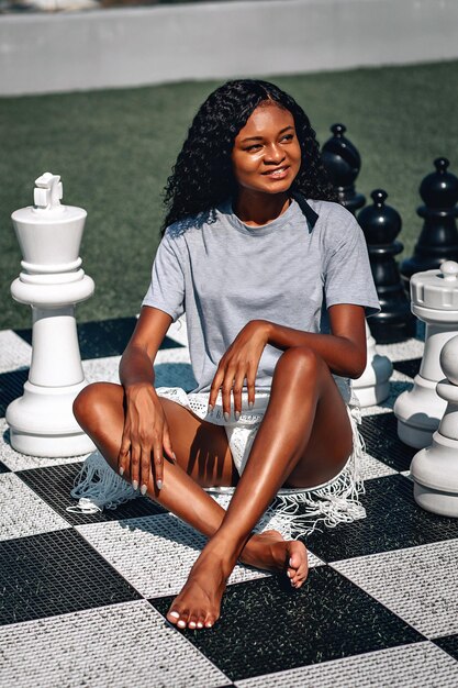 Smart African-American woman playing giant chess as she sits on the board with her legs crossed; glamourous intelligence concept.