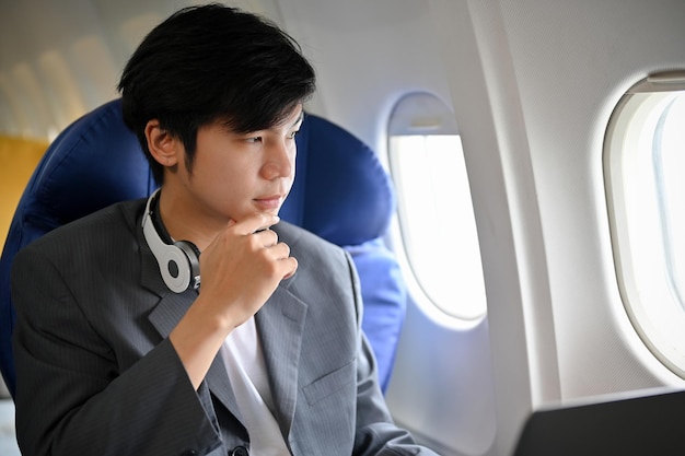 Smart asian businessman in formal suit is on the flight looking\
out the plane window