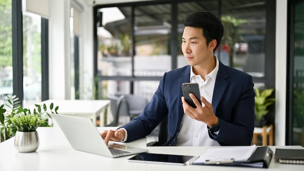 Умный азиатский бизнесмен держит свой смартфон и использует ноутбук за своим столом