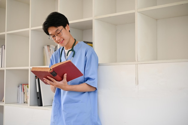 Smart Asian male medical student wearing glasses and reading a book while standing in library