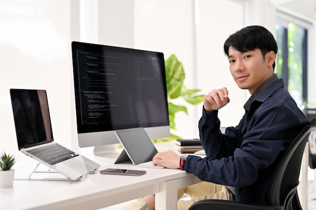 頭の良いアジア人男性のウェブ開発者が、現代のオフィスデスクに座り、笑顔でカメラを見ている