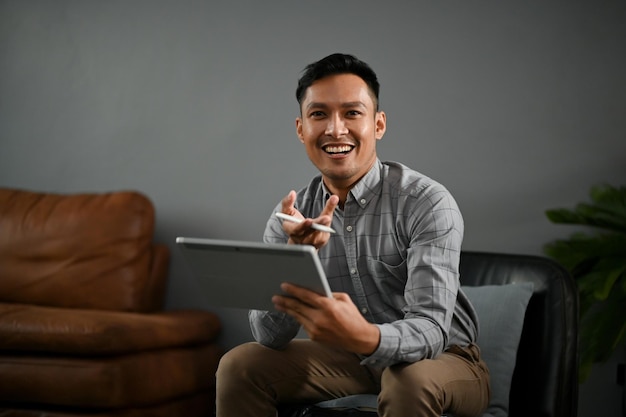 Smart Asian man in casual clothes sits on a black leather chair with his digital tablet