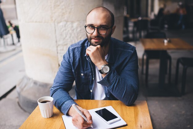 Smart bearded man in glasses writing at table and looking at camera