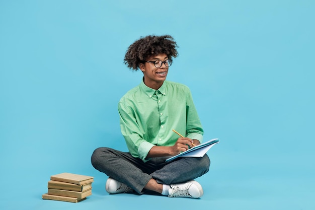 Smart black teen guy in glasses taking notes holding notepad and pen sitting with books nearby on