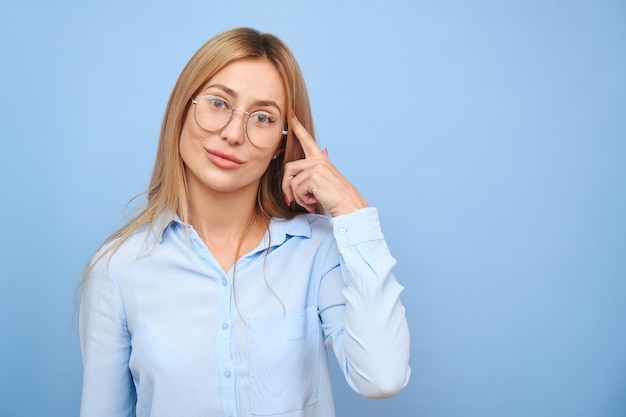 Smart blonde girl in business shirt and glasses touches her head thinks doubts makes decision isolated on blue studio background