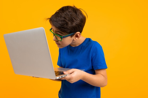 Smart boy in a blue T-shirt carefully typing on a laptop on a yellow studio background