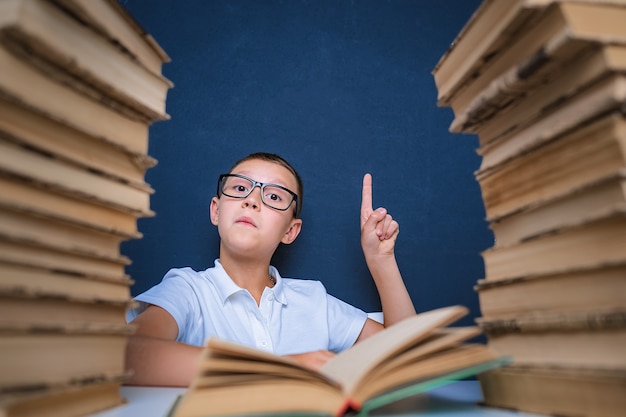 Smart boy in glasses sitting between two piles of books and look up, pointing finger.