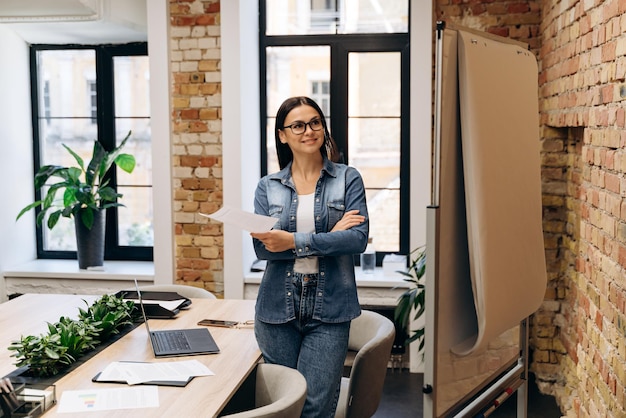 Smart brunette business woman standing near flipchart and thinking about presentation of new project in office interior