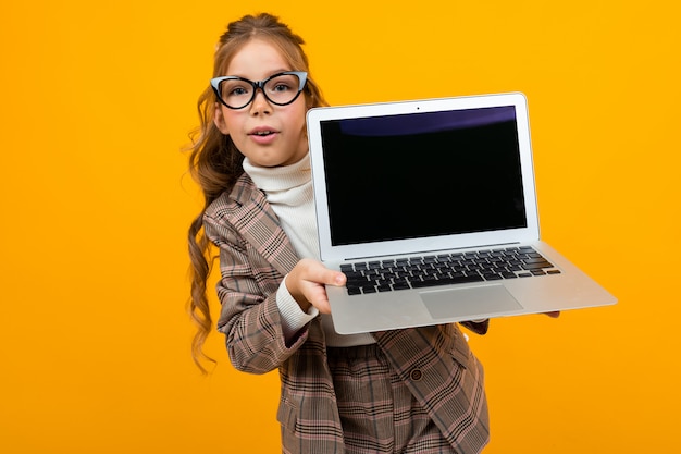 Smart Caucasian schoolgirl with glasses in a business suit and a jacket with a laptop with a layout in his hands explains the theory onn orange