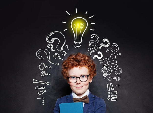 Photo smart child boy in glasses smiling and holding book on background with lightbulb