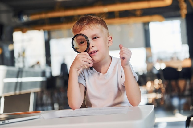 Smart child in casual clothes with laptop on table have fun with magnifying glass