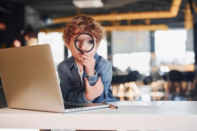 Foto il bambino intelligente in abiti casual con il laptop sul tavolo si diverte con la lente d'ingrandimento