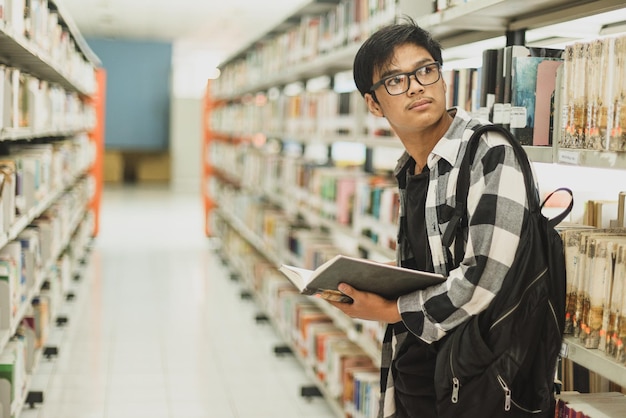 Smart college student standing near bookshelf in college library looking away while holding book