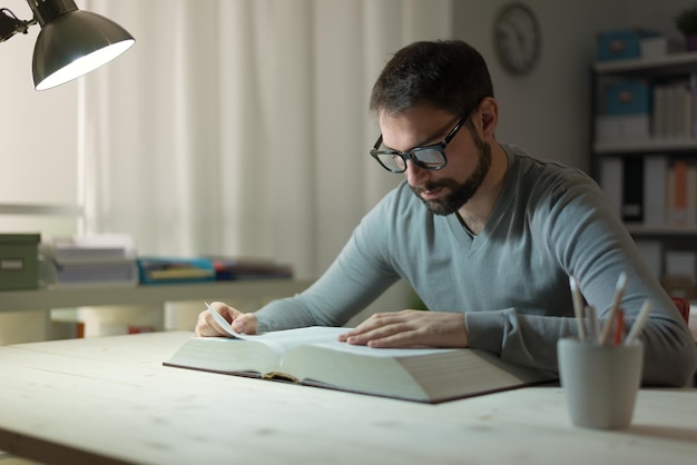 Photo smart confident young man studying late at night he is sitting at desk and reading a book knowledge and learning concept