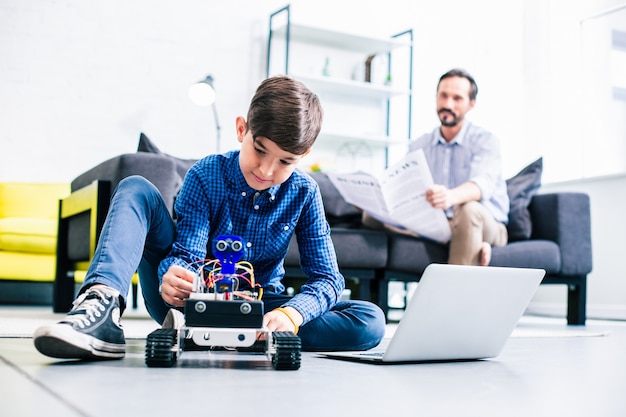 Smart delighted boy testing his robot while preparing for engineering classes