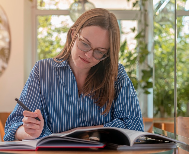 Smart female student in eyeglasses in modern cafe with books and workbooks studying in coffee house