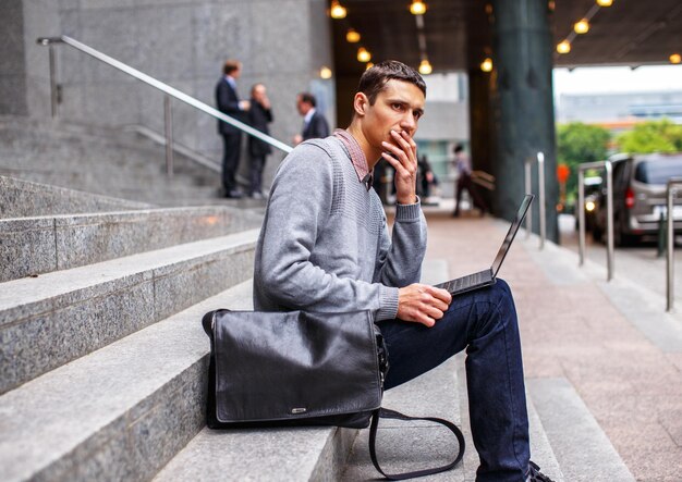 Smart guy in grey shirt and jeans sitting on stairs and working with laptop