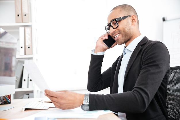 Smart happy usinessman using mobile phone and looking at documents in office