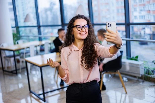 세련된 옷과 안경을 쓴 똑똑하고 행복한 젊은 비즈니스 여성은 사무실에서 일하고 스마트폰, 제스처, 미소로 고객과 이야기합니다. 직원들은 백그라운드에서 일하고 있습니다. 선택적 초점