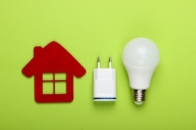 Smart house concept. Figurine of house and energy-saving light bulb with charger on green background. Top view
