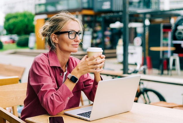 コーヒーショップに座ってコーヒーを飲みラップトップでリモートで作業している賢い現代の女性