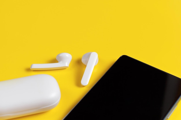Smart phone and white wireless earphones with the case top view on yellow background close up