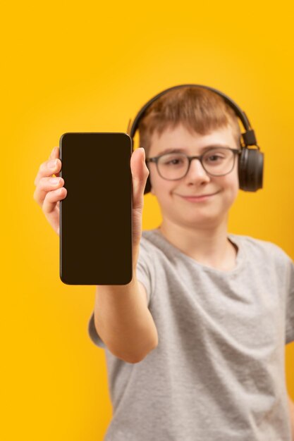 Smart phone with black blank screen in hands of teenage boy in headphones on yellow background Vertical frame Copy space