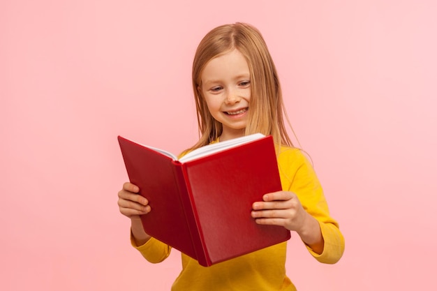 Smart positive cute little girl reading big red book and smiling happily learning homework being curious about school knowledge education concept indoor studio shot isolated on pink background