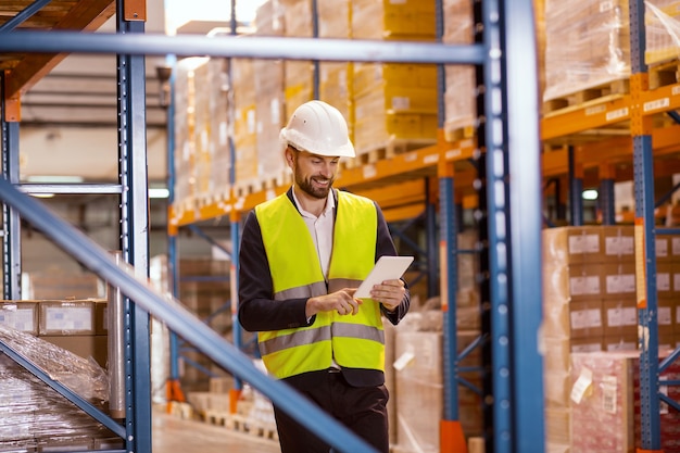 Smart positive man using a tablet while checking the working process in the warehouse