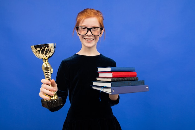 Ragazza adolescente dai capelli rossi intelligente in bicchieri con una pila di libri e una coppa della vittoria su uno sfondo blu studio.