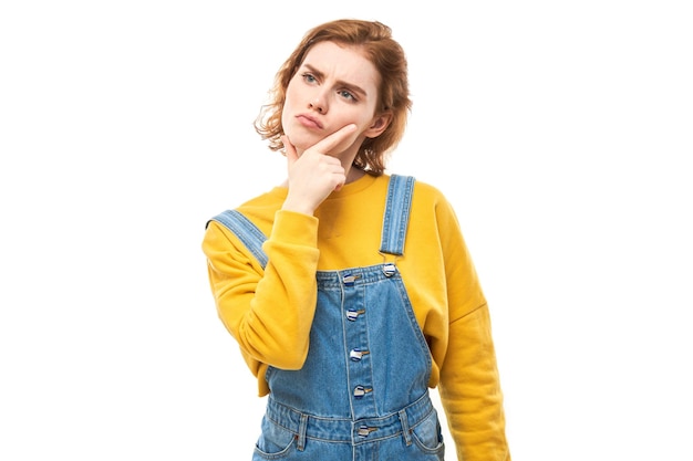 Smart redhead girl in casual holding chin thinks doubts makes decision isolated on white studio background