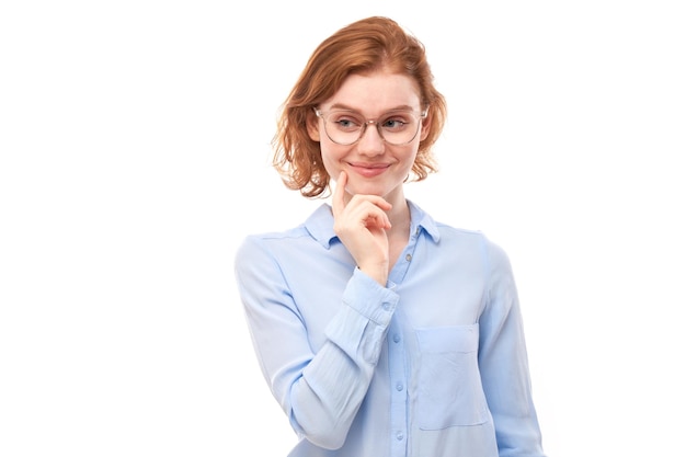 Smart redhead girl in glasses and business shirt holding chin thinks doubts makes decision isolated on white studio background