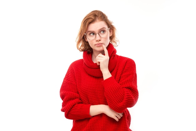 Smart redhead girl in red and glasses holding chin thinks doubts makes decision isolated on white studio background