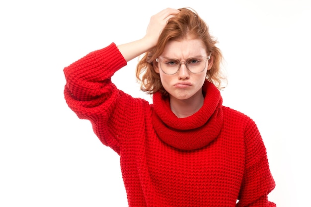 Smart redhead girl in red and glasses touching her head thinks doubts makes decision isolated on white studio background