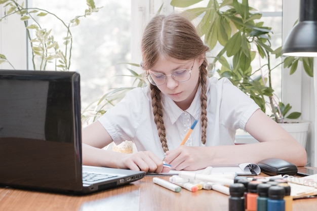 Smart schoolgirl does homework sitting at her desk writes with a pen in her writing notes and uses laptop. She learning language online, doing school tasks at home, listening to lecture