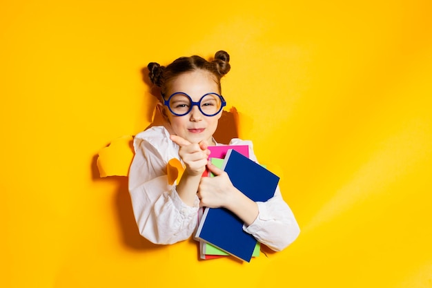 Photo smart schoolgirl looking through hole in yellow paper back to school little girl in eyeglasses holding books